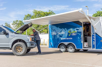 A mechanic works on a gray Ford pickup truck with the hood open, beside a 'Gilbert Ford Mobile Service' trailer.