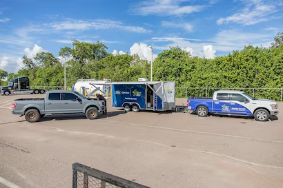 A grey Ford F-150 truck getting serviced by a technician from a blue Ford Mobile Service trailer, with another truck and trailer in the background.